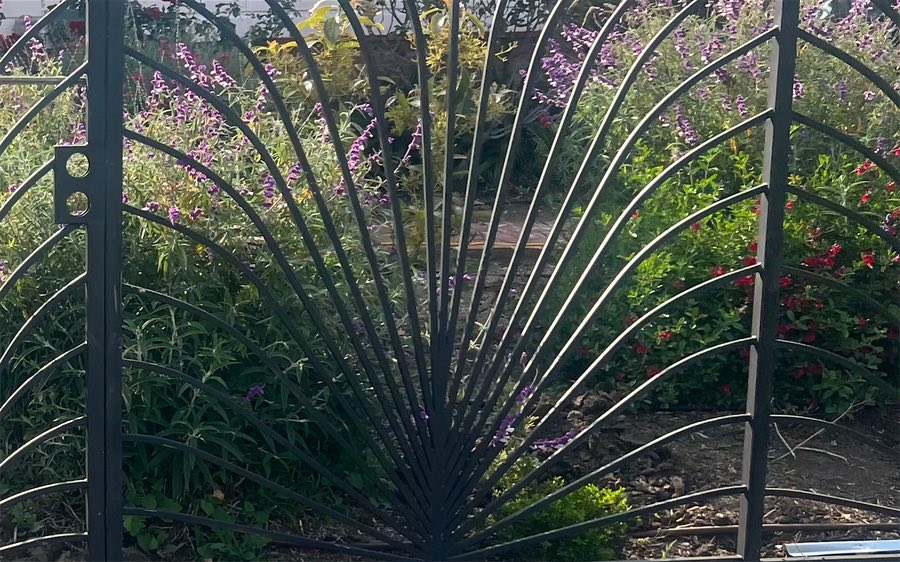 Close-up of an ornamental fence whose bars fan out from a single point, in front of a flower garden
