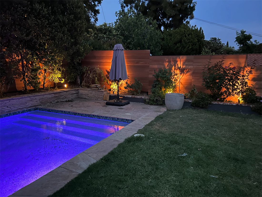 A backyard at dusk: a horizontal slat fence washed with landscape lighting, behind a lit pool and lawn