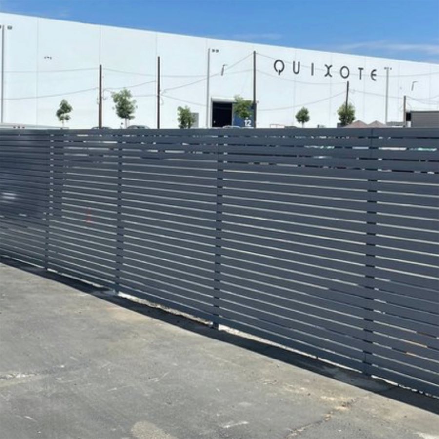 A long horizontal slat fence along the parking lot of a commercial building bearing the Quixote sign