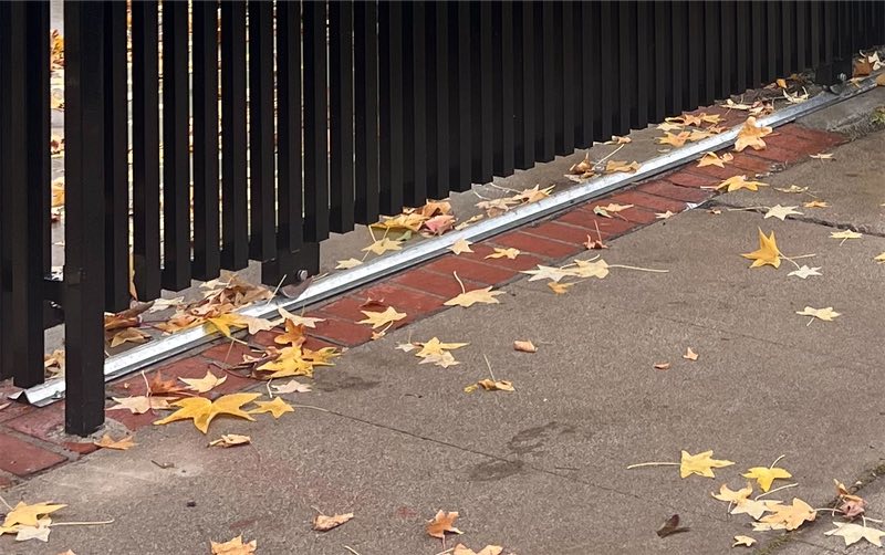 The foot of a black sliding gate and its steel track along a brick edge, autumn leaves on the sidewalk