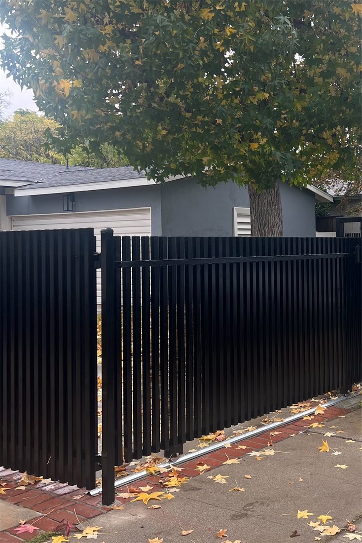 A black vertical bar fence and sliding gate on its track, under a large tree in autumn