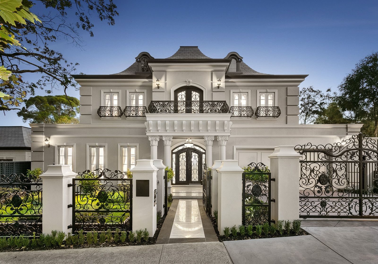 A classical two-storey home at dusk behind ornate wrought-iron gates, fencing and balconies